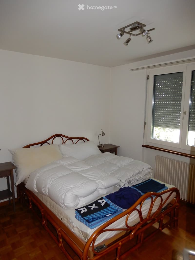 Cozy bedroom with a wooden bed, white bedding, nightstand, and a lamp. The room features wooden flooring and a radiator under the window.