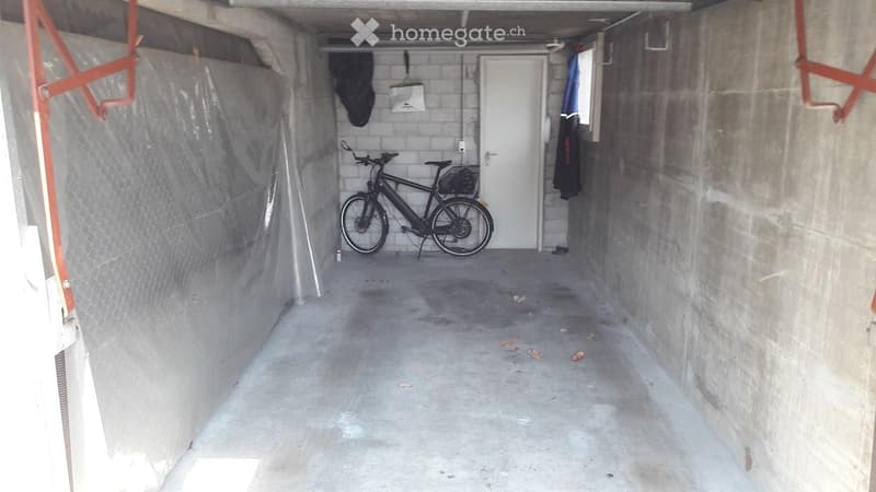 Concrete garage with a bicycle parked inside, a white door at the back, and some protective coverings on the walls.