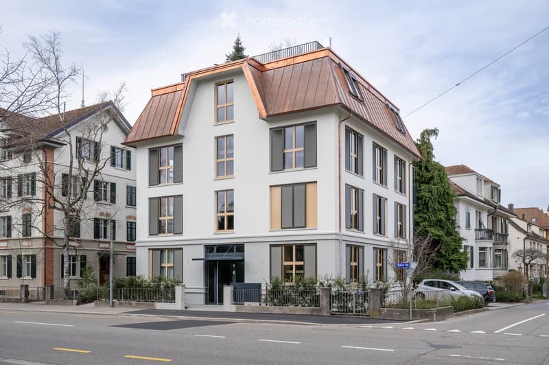 Modern apartment building, copper roof, white walls, large windows, fenced yard, and a view of the street.