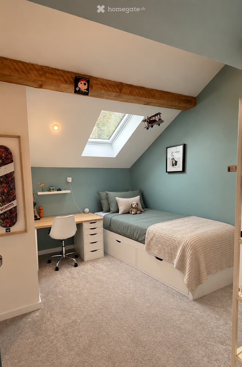 Cozy attic bedroom with a slanted wooden ceiling, skylight window, built-in bed with storage drawers, desk with chair, and decorative items including framed pictures and a toy airplane.