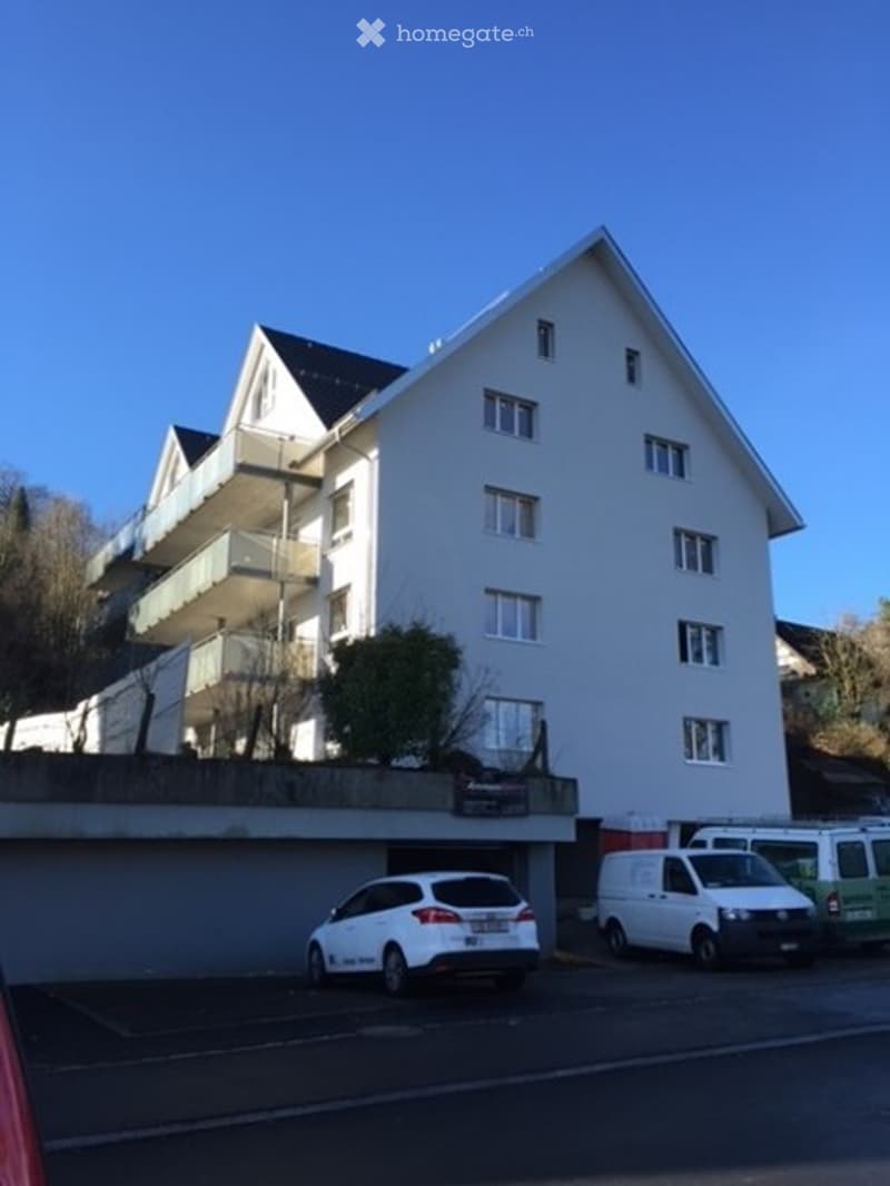 Multi-story residential building with white exterior, multiple balconies, and a parking area with several cars.