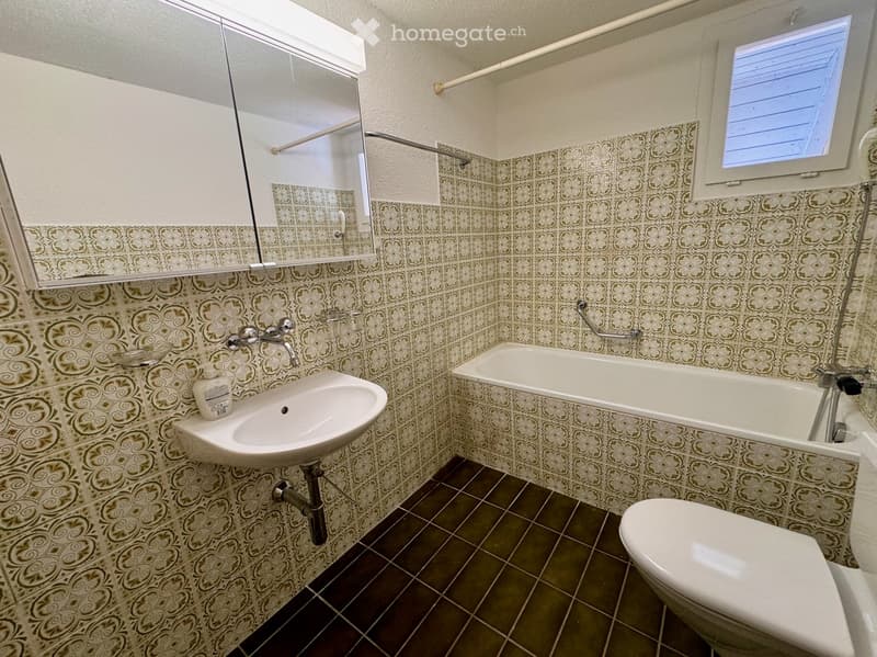 Bathroom with floral patterned tiles, bathtub, toilet, sink, mirror, and a small window.