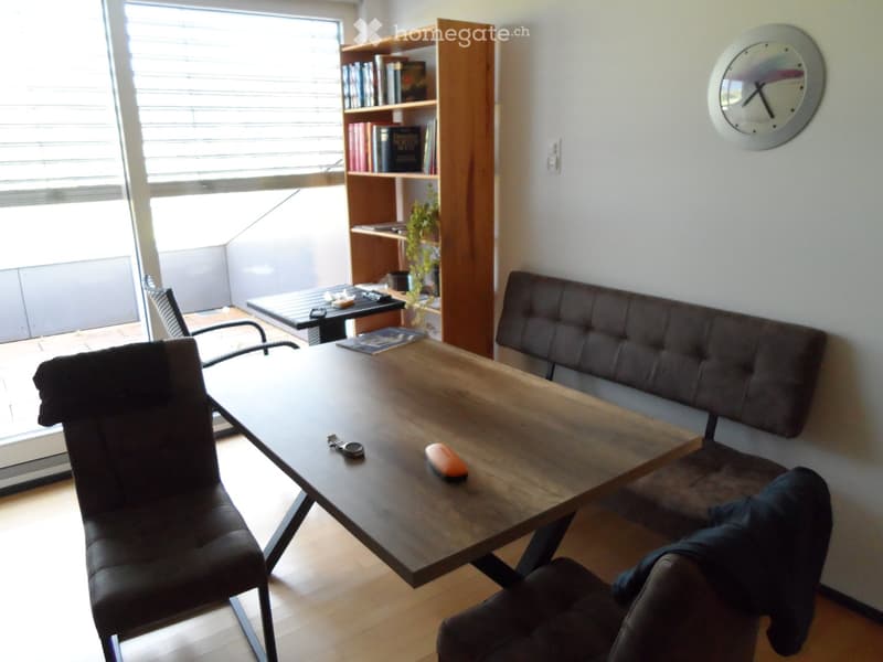A modern living room with a dark wooden table, two black chairs, a grey sofa, a bookshelf filled with books, and a wall-mounted clock.