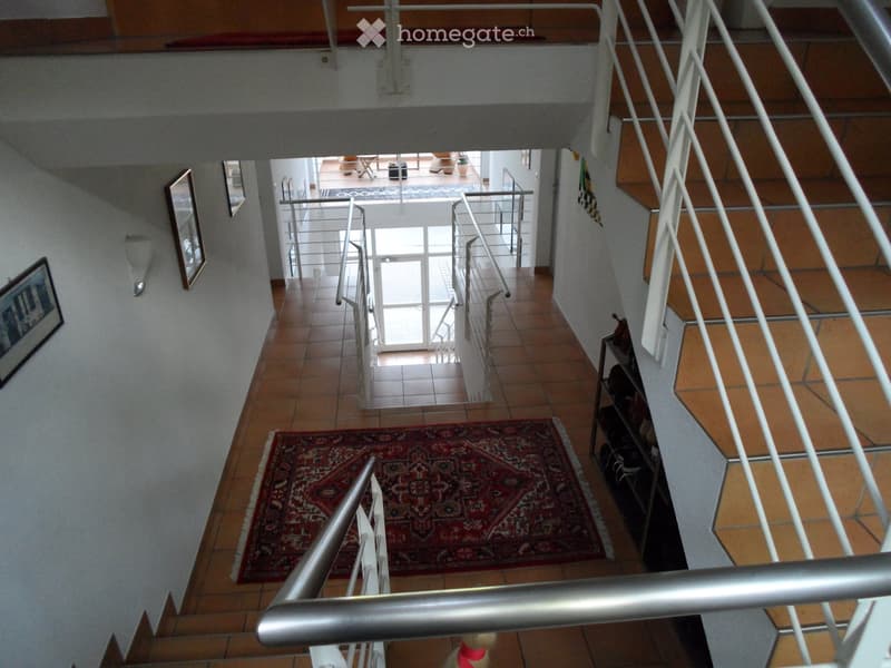 Spacious hallway with tiled flooring, metal railings, and a red patterned rug on the stairs. The hallway leads to an open area with large windows and a balcony.