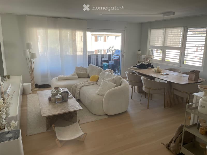 Modern living room with a curved sofa, coffee table, dining table, chairs, cat tree, and a small fan. Large windows and a sliding glass door lead to a balcony.