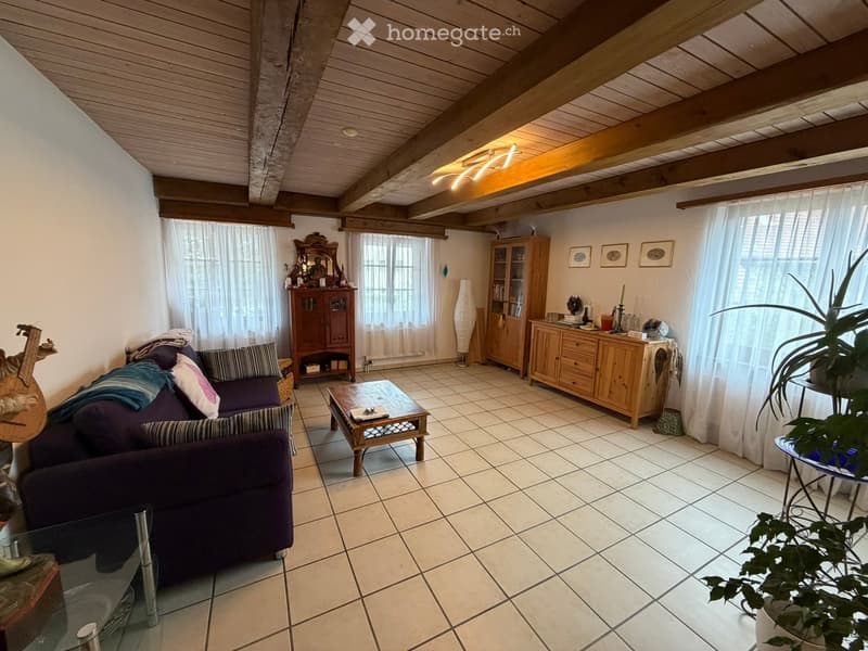 Living room with exposed wooden beams, tiled floor, and large windows. Features a purple sofa with pillows, a wooden coffee table, a cabinet with glass doors, a mirror, and a guitar leaning against the wall.