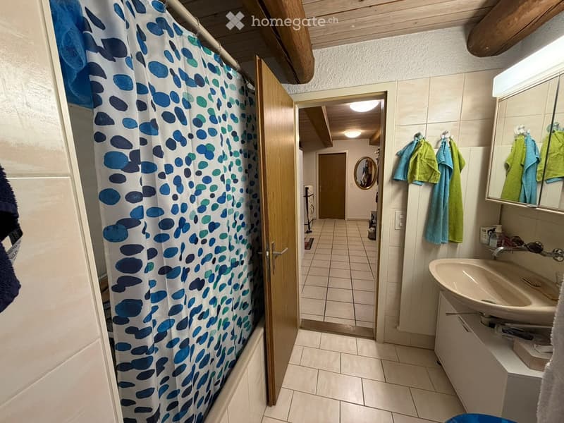 Bathroom with a sink, mirror, towels, and a shower curtain. The room has tiled floors and walls, with a wooden door leading to another area.