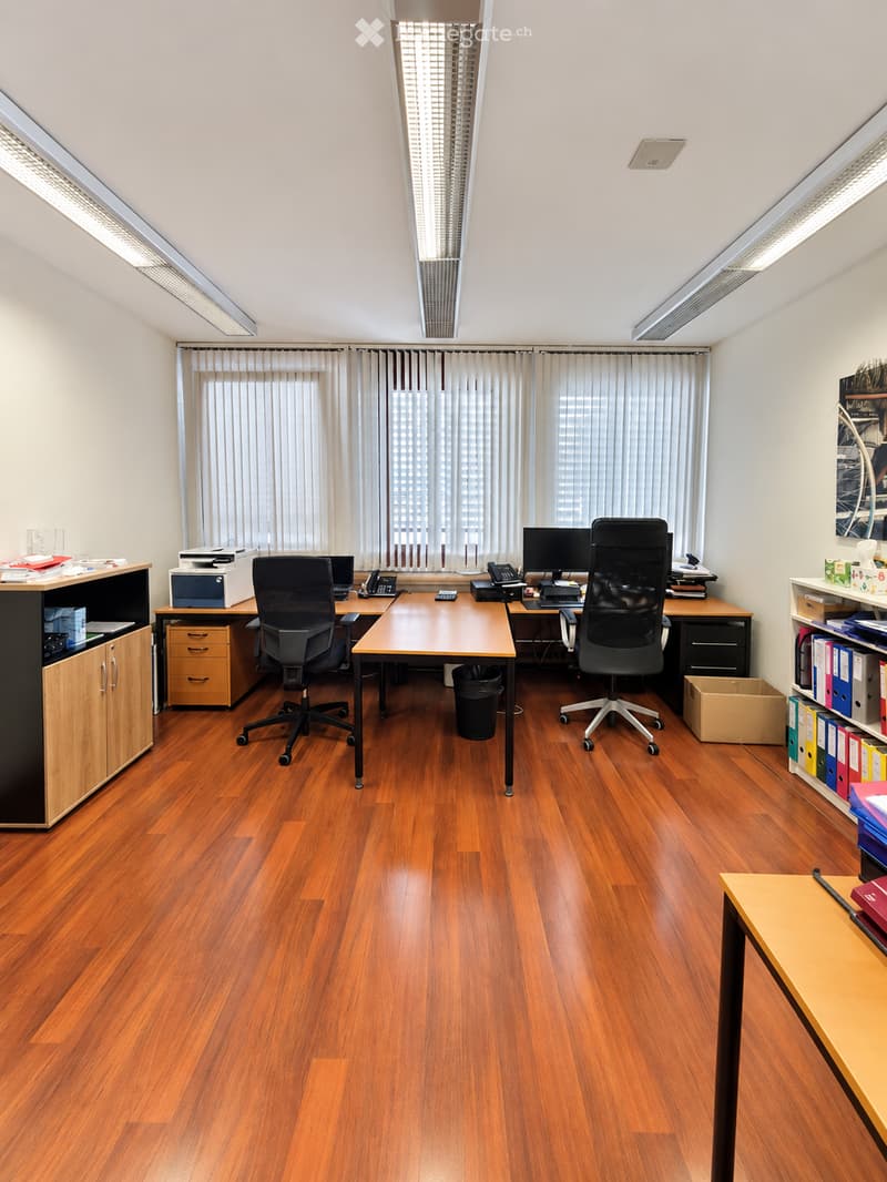 Office space with wooden flooring, large windows with blinds, two desks with office chairs, computers, printers, and filing cabinets.