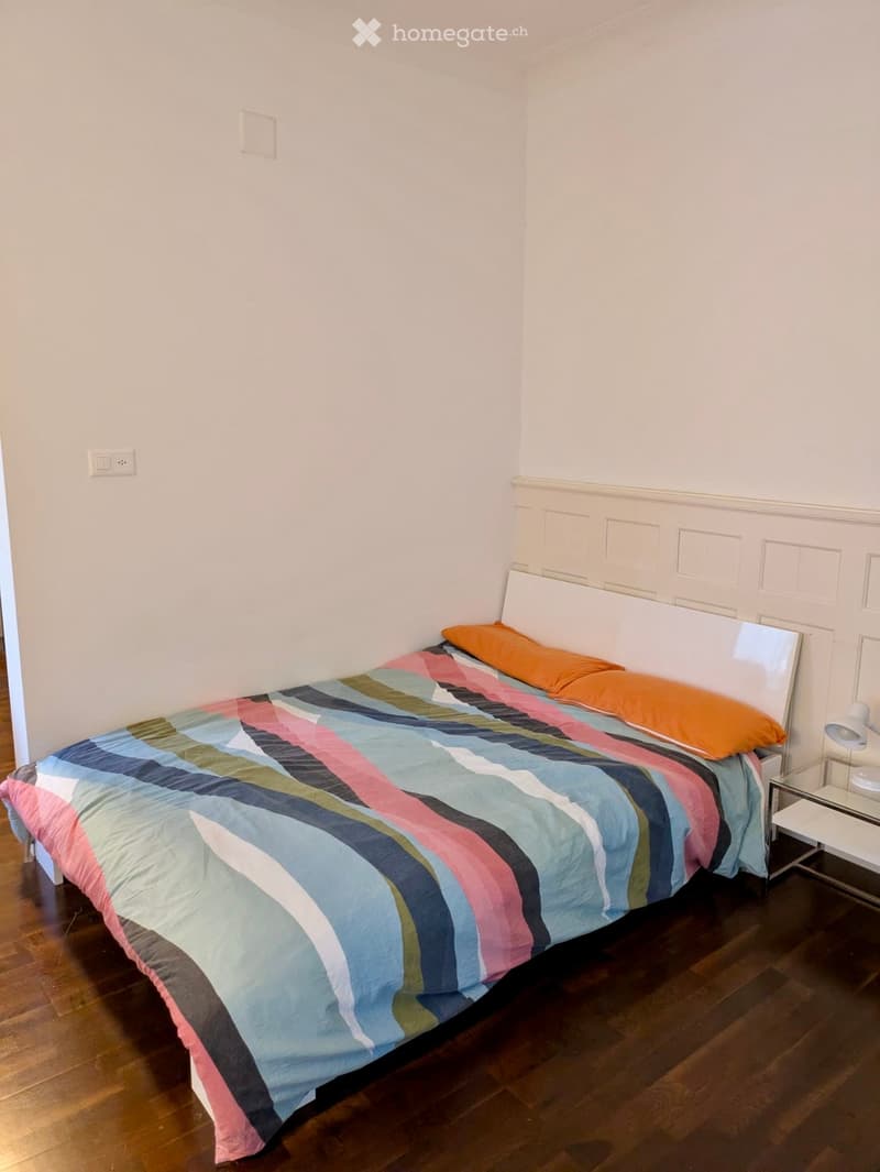 A small bedroom with a single bed, colorful striped bedspread, orange pillows, a white headboard, and a glass nightstand with a lamp.