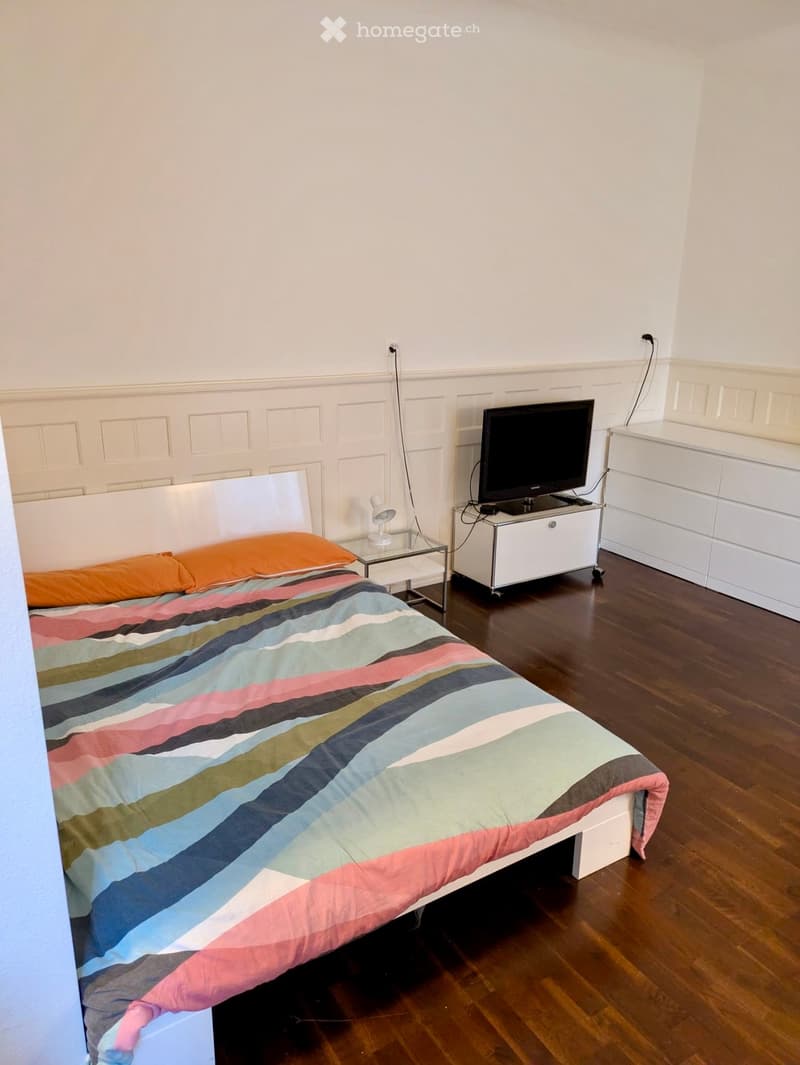 A modern bedroom with a wooden floor, a white bed with a colorful striped duvet, a television on a white stand, a nightstand with a lamp, and a white dresser.