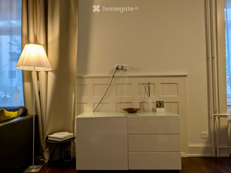 Modern, minimalist room with a sleek white drawer unit, a glass-topped table, a floor lamp, a sofa with a yellow pillow, and decorative items on the table.