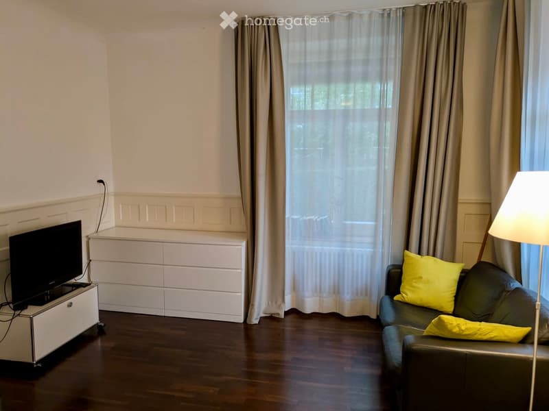 Modern living room with dark wooden flooring, a grey leather sofa with yellow pillows, a flat-screen TV on a white stand, a white cabinet, and a floor lamp near the window.