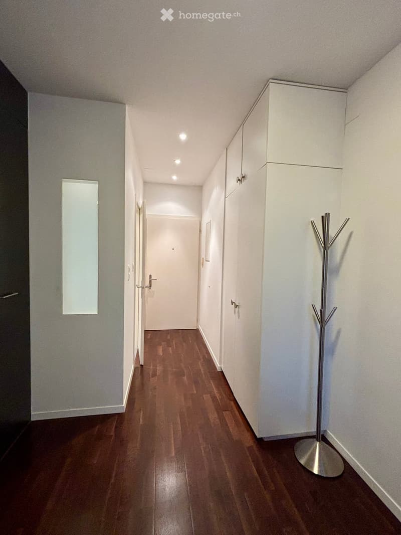 A narrow hallway with wooden flooring, white walls, and built-in white cabinets. A coat rack is positioned on the right side.