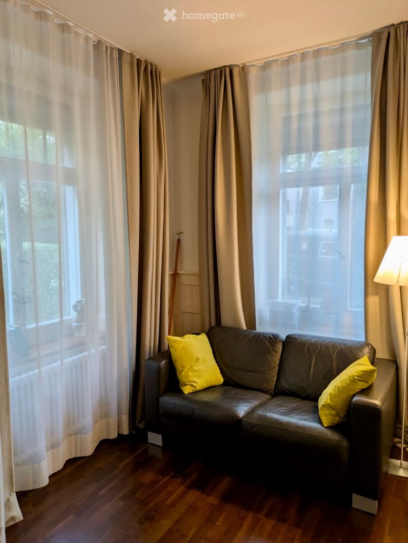 Living room with a dark grey leather sofa, yellow pillows, and a floor lamp near large windows with sheer curtains.