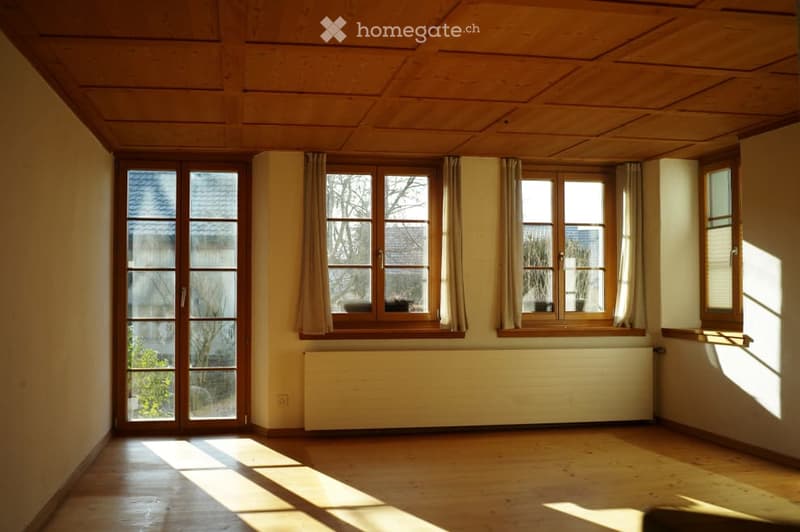 Empty room with wooden flooring, large windows with curtains, and a radiator. No furniture or equipment present.
