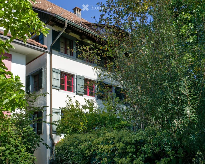 A two-story white house with red-framed windows and green shutters, partially obscured by lush greenery and trees.