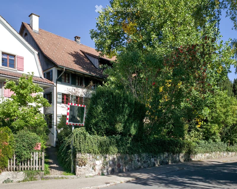 Two-story house with a red-tiled roof, white walls, and multiple windows, surrounded by lush greenery and trees.