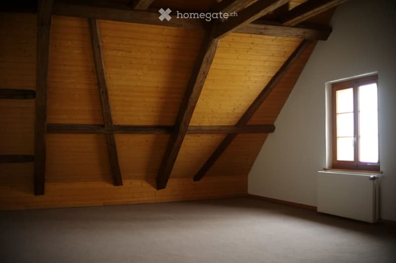 Empty attic room with wooden beams, exposed wooden walls, and a single window with a radiator below it.