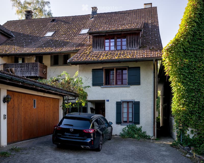 Two-story house with a tiled roof, multiple windows, and a wooden garage door. A black car is parked in front of the house.