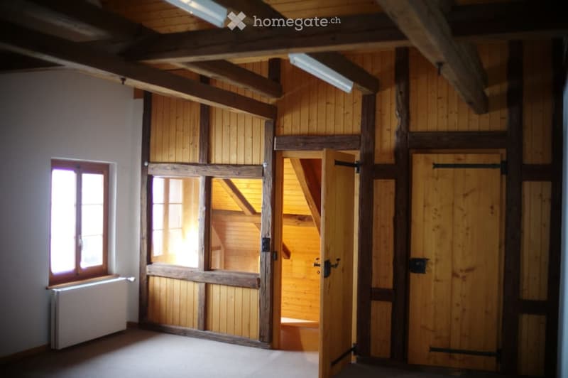 Empty room with wooden walls, exposed wooden beams, and large wooden doors. Natural light from windows illuminates the space.
