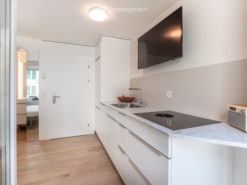 Modern kitchen with white cabinets, marble countertop, induction stove, sink, and a bowl on the counter. A radio is placed on the counter, and a TV is mounted on the wall.