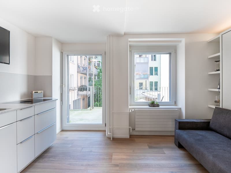 Modern, open-plan living and dining area with light wood flooring, white kitchen cabinets, a grey sofa, and large windows offering a view of a balcony and neighboring buildings.