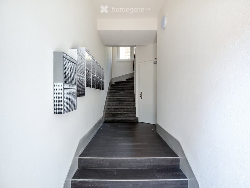 Modern hallway with a staircase leading upwards, white walls, dark tiled floor, and a white door at the end.