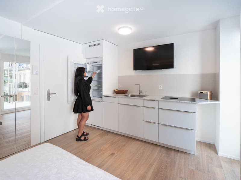 Modern kitchen with white cabinets, a stainless steel sink, a refrigerator, a countertop, a bowl, a radio, and a television mounted on the wall.