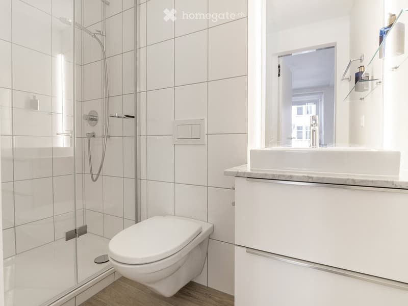 Modern bathroom with white tiles, a glass-enclosed shower, a toilet, a sink with a mirror, and a cabinet.