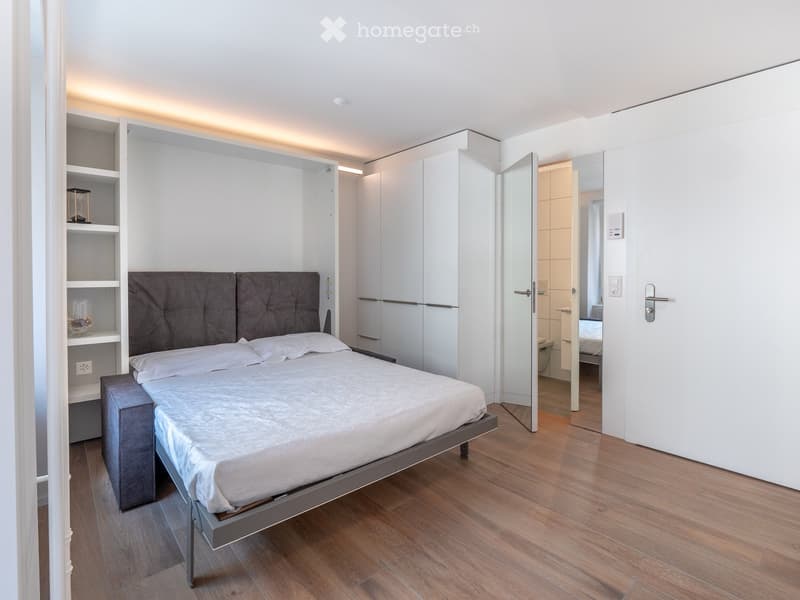 Modern bedroom with a large bed, grey headboard, built-in wardrobe, and shelves. The room features wooden flooring and white walls.