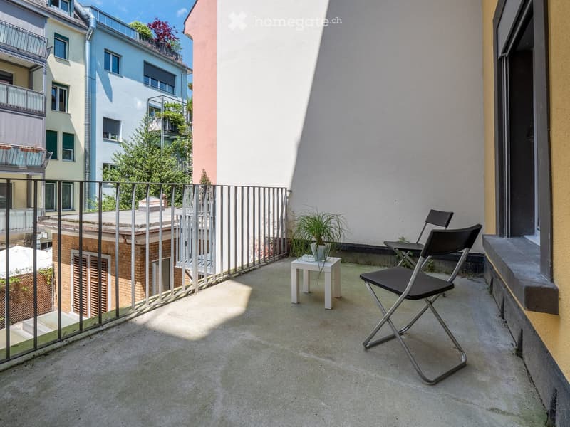 Balcony with a metal railing, concrete floor, two black folding chairs, a small white table with a potted plant, and a view of neighboring buildings and greenery.