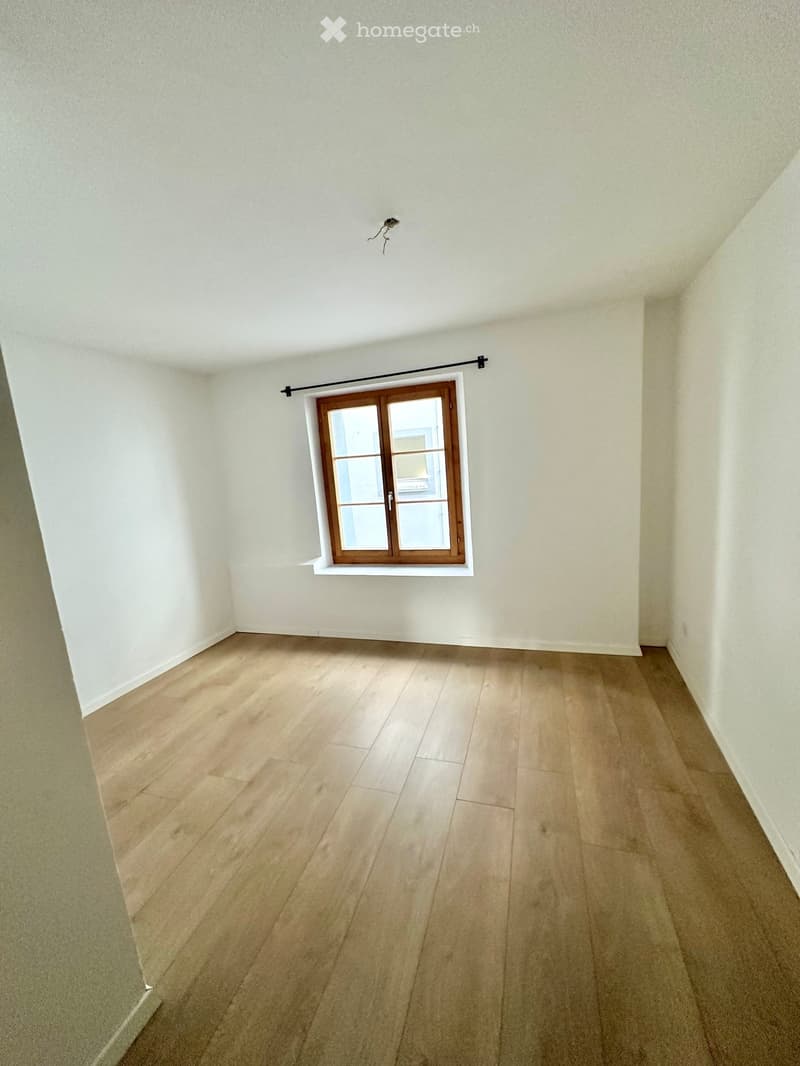 Empty room with light wooden flooring and white walls, featuring a single window with wooden frame and a black curtain rod above it.