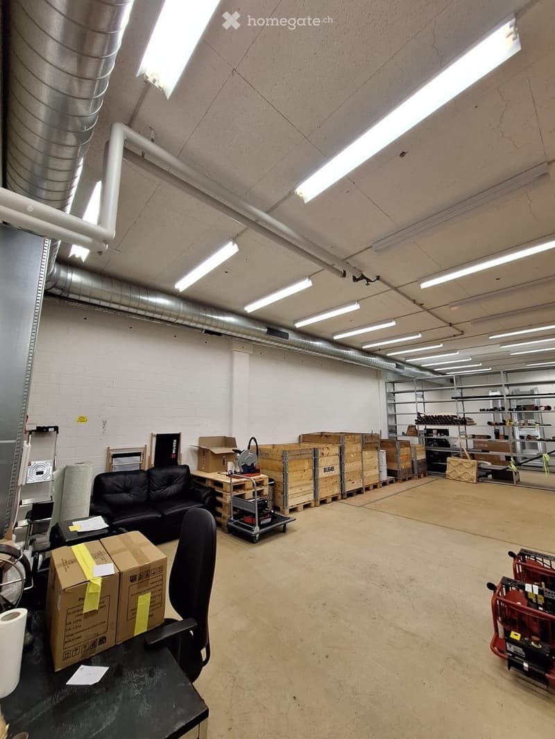 Large industrial-style room with exposed pipes and lighting, containing a black sofa, office chair, table, shelves, and storage boxes.