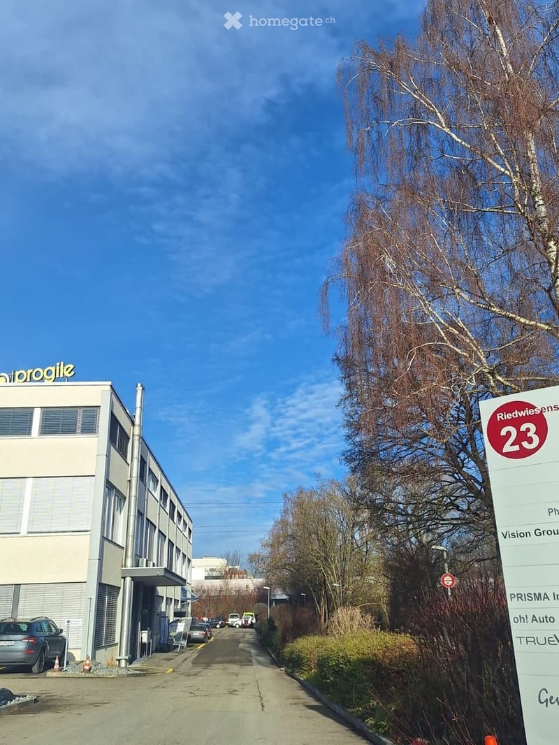 Modern multi-story building with signage for "Progile" and "Riedwiesen23" visible, surrounded by trees and parked cars.