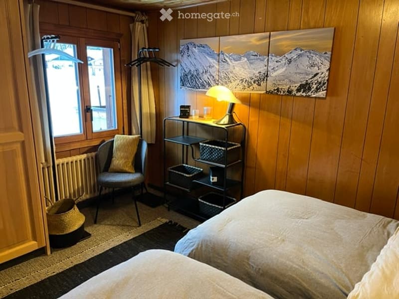 Cozy bedroom with wooden walls, a double bed, a chair with a pillow, a small table with a lamp, and a picture frame on the wall.