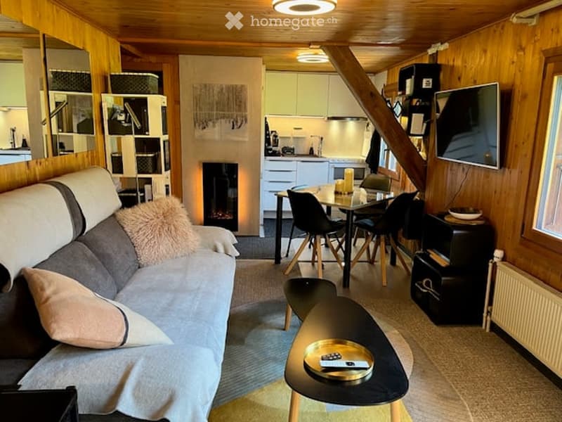 Cozy, open-plan living and dining area with wooden ceilings, a fireplace, a grey sofa with throw pillows, a coffee table, a dining table with chairs, and a television mounted on the wall. The kitchen area features white cabinets and modern appliances.
