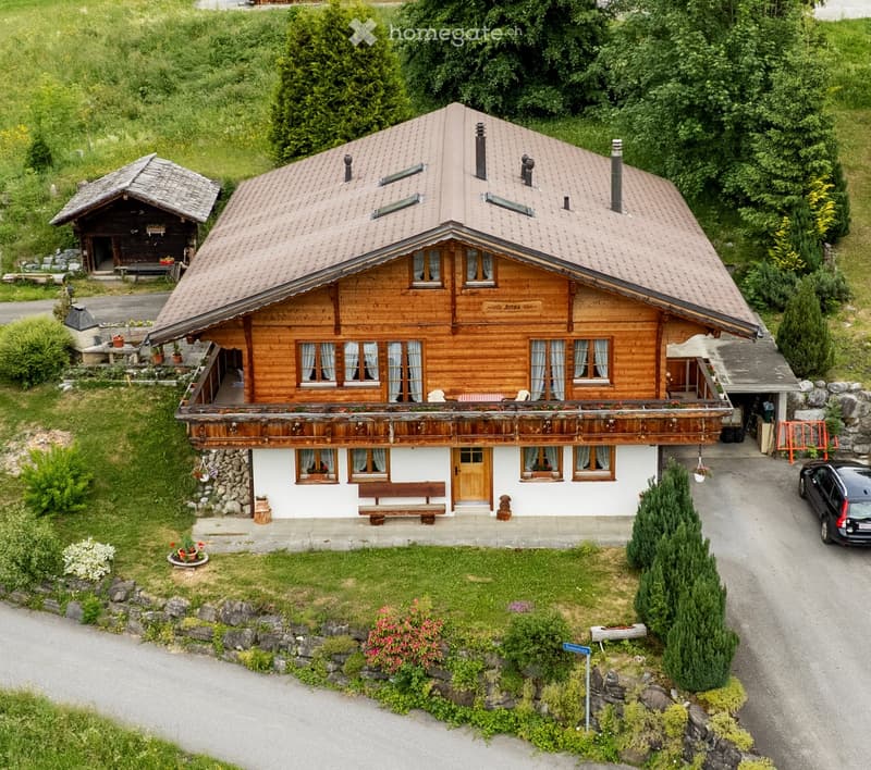 A two-story wooden house with a sloped roof, multiple windows, and a balcony. The house is surrounded by greenery and has a small garden area with plants and a bench.