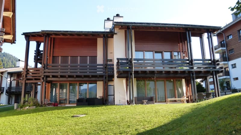 Modern multi-story building with wooden balconies and large windows, surrounded by a well-maintained lawn.
