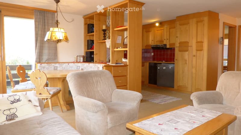 A cozy living and dining area with a sofa, armchair, coffee table, dining table, and chairs. The space features wooden cabinets, a dishwasher, and a shelf with decorative items.