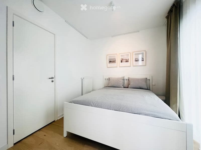 A small, modern bedroom with a white bed, grey bedding, two pillows, a nightstand, and three picture frames on the wall.