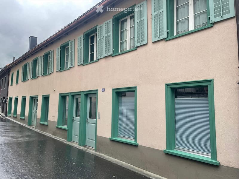 A two-story building with light beige walls, green shutters, and multiple windows. The ground floor has closed shutters and doors, while the upper floor features open windows with visible interior elements.