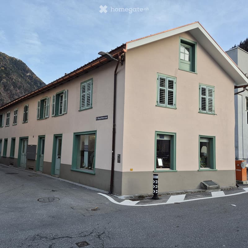 Two-story building with light pink facade, green shutters, and multiple windows. The building has a sloped roof and is situated on a street with a visible mountain in the background.