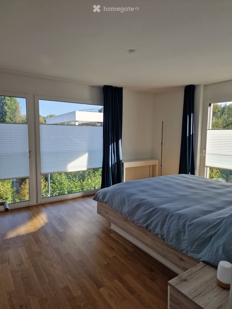 Modern bedroom with wooden flooring, large windows with blinds and dark curtains, a neatly made bed, and a nightstand with a lamp.