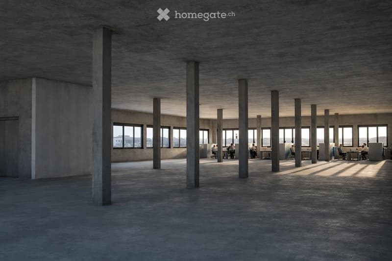 Open-plan office space with concrete floors and pillars, large windows, and a few desks and chairs arranged in groups.