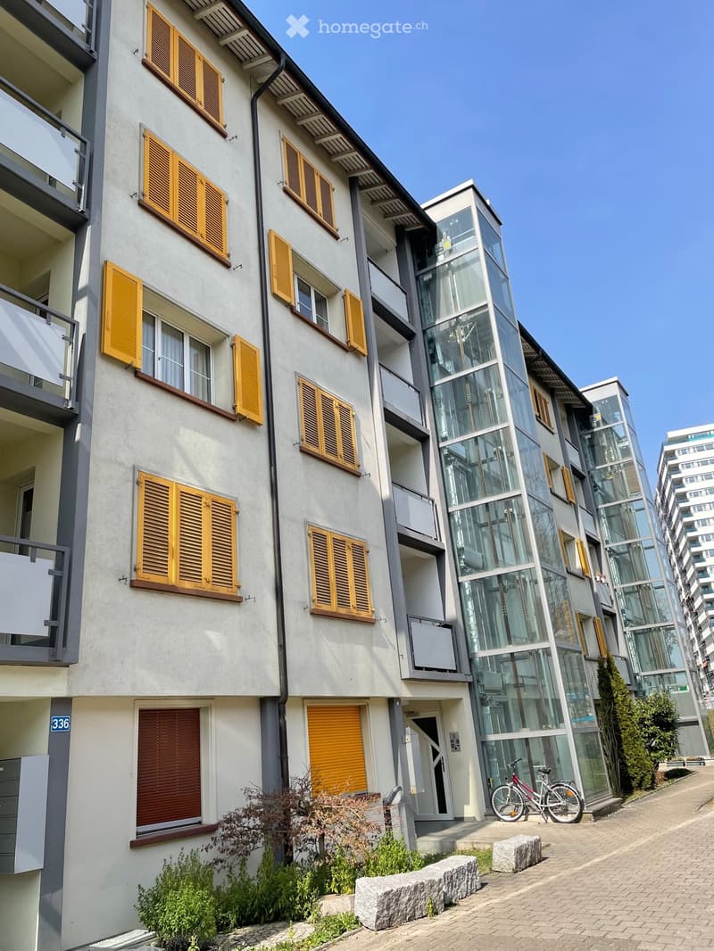 Modern multi-story building with white facade, yellow shutters, and glass balconies. Features include a bicycle parked near the entrance and small landscaped area with shrubs and stone benches.