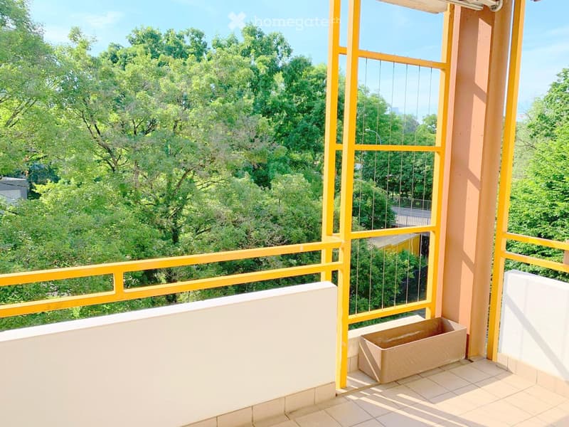 Balcony with yellow railings, tiled floor, and a small planter, overlooking a lush green area with trees.
