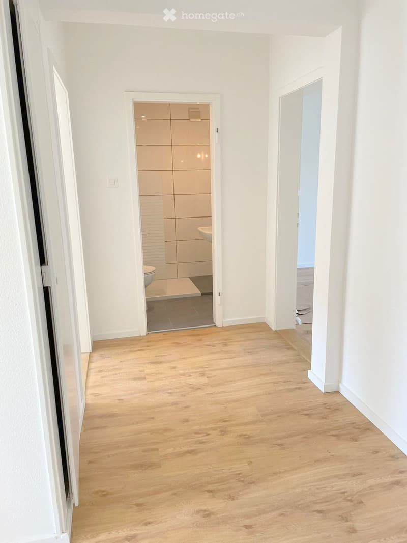 A clean, modern hallway with light wooden flooring and white walls. The hallway features a partially open door leading to a bathroom with light-colored tiles and a visible toilet, and another door leading to another room.