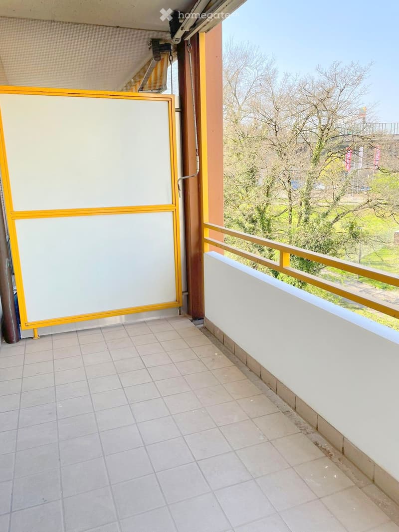Balcony with tiled floor, white walls, yellow-framed sliding doors, and a view of trees and a road.