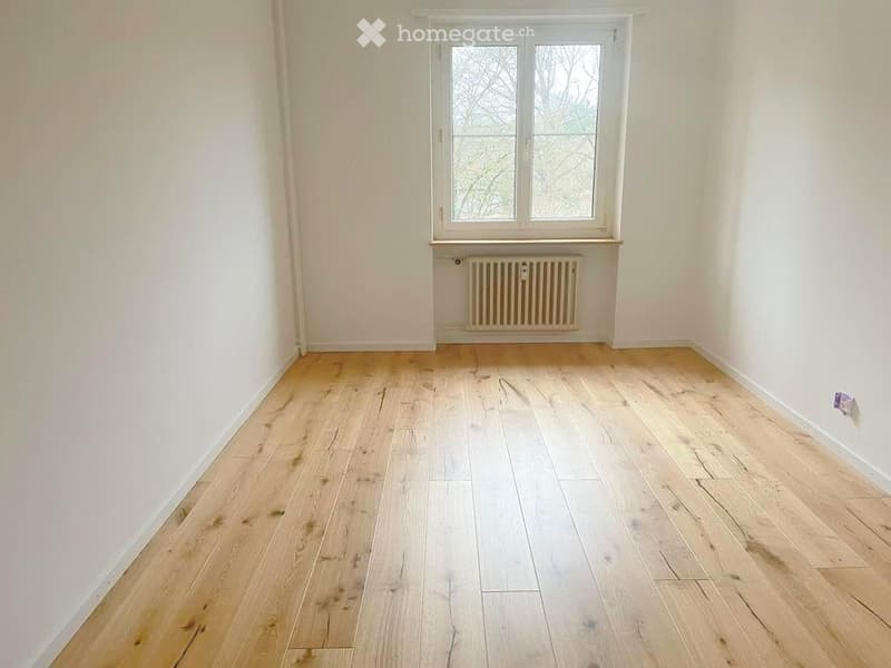 Empty room with light wooden flooring, white walls, and a single window with a radiator below it.