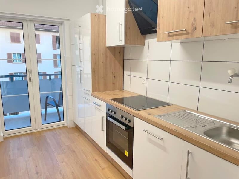 Modern kitchen with white cabinets, wooden countertops, induction stove, oven, and a sink. A sliding glass door leads to a balcony.